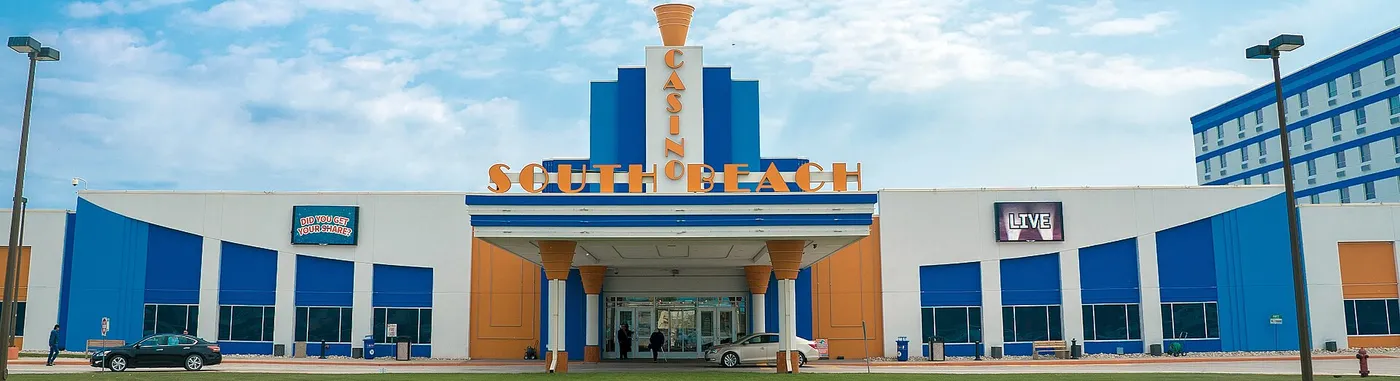 Photograph of the front of South Beach Casino & Resort: a white, blue and orange Art-deco entrance under a blue sky, with the hotel wing on the right.